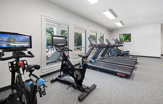 A room with exercise equipment and a TV screen showing a landscape Outside view of pool and clubhouse with pool furniture at The Palms at Chatham apartments in Savannah GA,