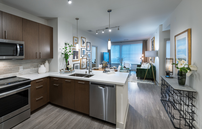 Open-concept kitchen, dining, and living space with ash-gray wood-style floors, dark modern cabinets, stainless steel appliances, white quartz countertops, and a spacious living area with a large window.