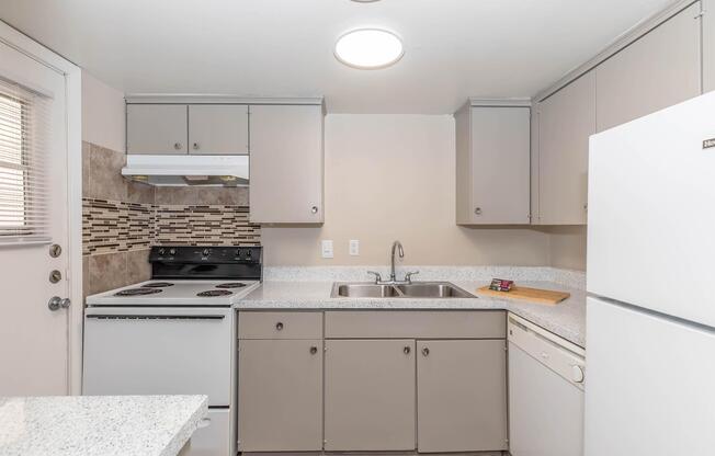 A modern kitchen featuring light gray cabinets, white appliances, a tile backsplash, a double sink, and a granite countertop. The kitchen includes a stove, refrigerator, and dishwasher, with natural light coming through a window and door.