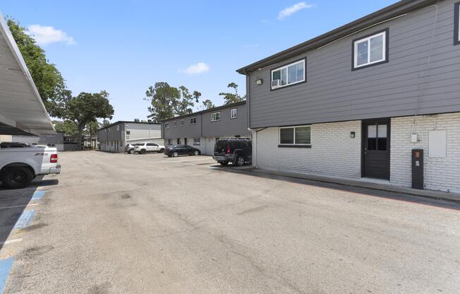 A view of an apartment complex featuring two-story buildings. A partially paved parking lot is visible, with several parked vehicles including trucks and sedans. The area is well-lit with clear skies and trees in the background, creating a serene suburban environment.