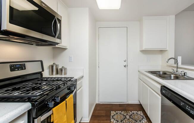 Modern kitchen featuring stainless steel appliances, including a microwave and gas stove. The white cabinetry complements the counters. A door is visible, leading outside, and a patterned rug adds a decorative touch to the floor. The space is well-lit and organized.