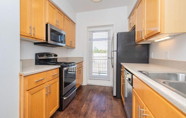 A modern kitchen featuring wooden cabinets, stainless steel appliances, including a microwave, oven, and refrigerator. The countertop is light-colored, and there's a double sink. A door leads to an outdoor space with blinds on a nearby window, allowing natural light into the room.