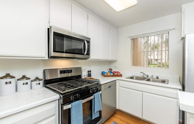 Modern kitchen featuring white cabinetry, stainless steel appliances including a microwave and stove, a double sink, and a window with blinds. A countertop with cooking utensils and containers is visible, along with light wood flooring.