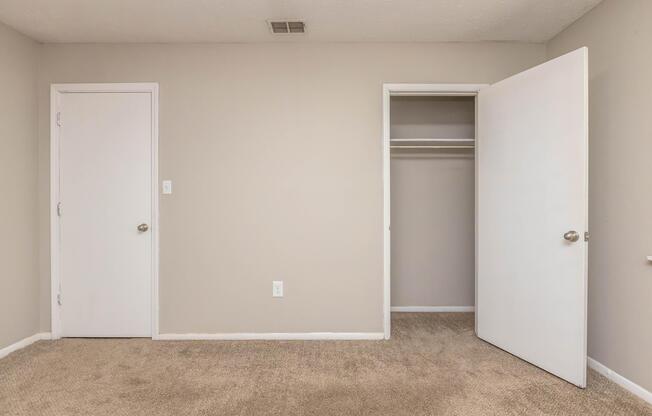 A minimalistic room with beige walls and carpet. On the left, there's a closed white door. On the right, a partially open door reveals a small closet with no items inside. The space appears bright and uncluttered, suggesting a neutral setting suitable for various purposes.