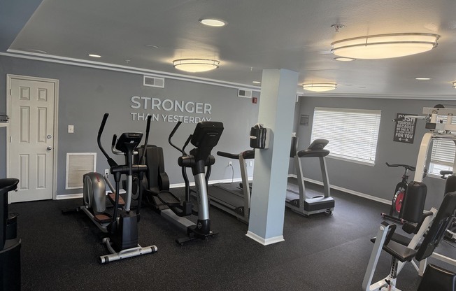A well-lit gym with exercise equipment, including elliptical machines and stationary bikes. The walls are painted light gray, and motivational signage reads "Stronger Than Yesterday." There are large windows allowing natural light, and the floor is covered with black rubber flooring for safety.
