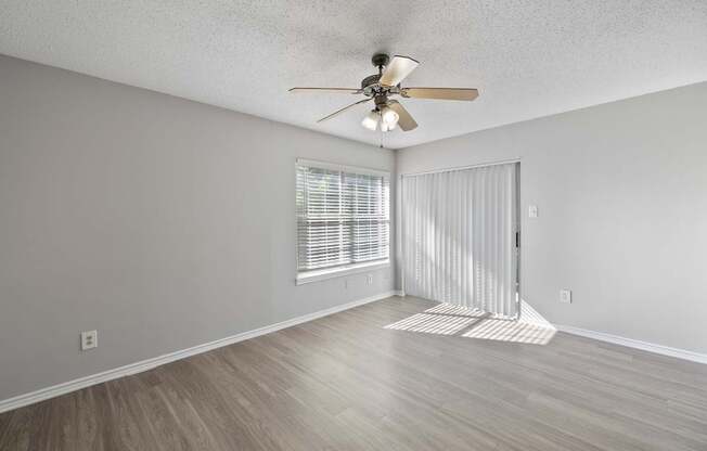 A room with a ceiling fan and light, wooden floors, and a window with blinds.