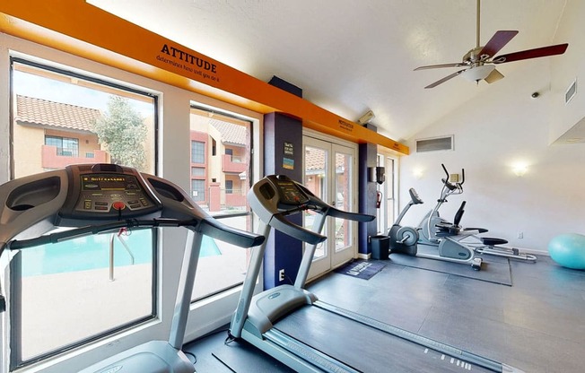 A bright fitness center here at Aspire Pinnacle Park featuring treadmills, elliptical machines, and cardio equipment positioned near large windows overlooking the pool area. Vaulted ceilings with a ceiling fan, motivational wall accents, and durable flooring create an energizing space designed for convenient, comfortable workouts close to home.