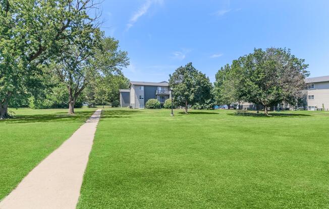 A sunny park scene featuring a well-maintained green lawn, lined with trees on either side. A paved walkway runs through the grassy area, leading toward a residential building in the background. The sky is clear and blue, enhancing the serene outdoor atmosphere.