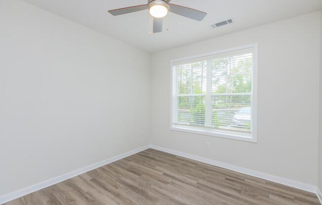A clean, empty room with light-colored walls and a wooden floor. A ceiling fan is visible, and a large window with white blinds allows natural light to enter, showcasing a view of greenery outside. The room is minimally furnished and inviting, suitable for various uses.