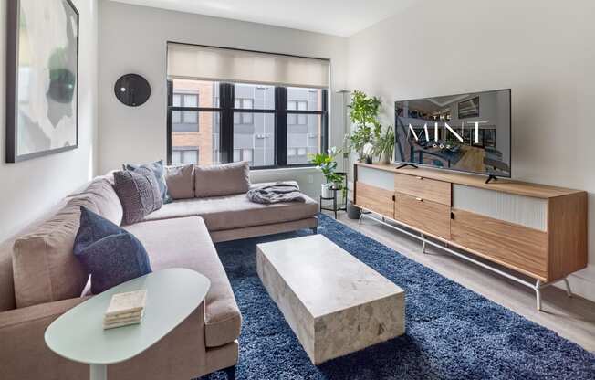 Comfortable living room at The Mint Rahway with a sectional sofa, modern decor, and a large window, providing a welcoming space for luxury apartment rentals in Rahway, NJ.