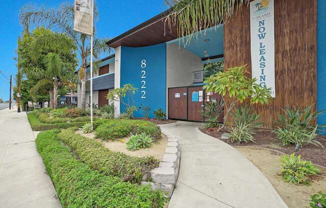 A building with a blue wall and a sign that says "NOW LEASING" is surrounded by greenery.
