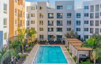 A swimming pool surrounded by lounge chairs and palm trees in front of apartment buildings.