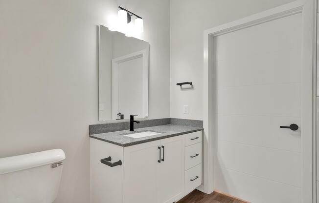 A modern bathroom featuring a white toilet, a sleek gray countertop with a rectangular sink, a large mirror above the sink, minimalistic black faucet and handles, and a light fixture with multiple bulbs. The wall is painted in a light color, and the door is a simple white design. Wooden flooring completes the look.